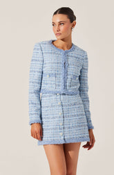 A woman is wearing a matching light blue tweed jacket and skirt set. The jacket is cropped with long sleeves, a round neckline, and decorative buttons on the front and on the sides. The skirt is high-waisted with a row of buttons down the front. The fabric has a textured, woven appearance with a mix of light blue, white, and darker blue threads. The woman has dark hair pulled back and is accessorized with hoop earrings.